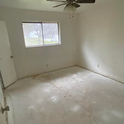 An empty bedroom with white walls, a ceiling fan, a window, and an unfinished floor with a visible crack.