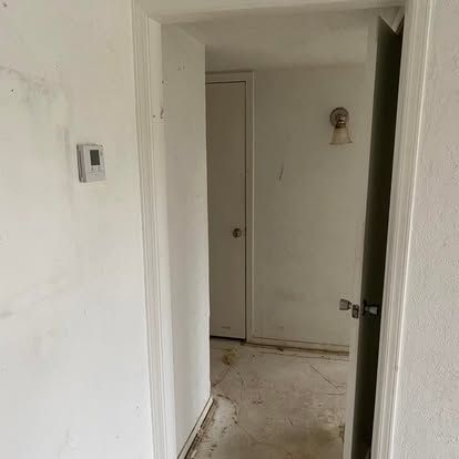 A view through a white doorway showing a hallway with a closed door, a wall-mounted light fixture, and an unfinished floor.