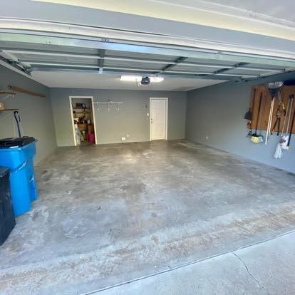An empty, gray-walled two-car garage with concrete floors, a doorway, a storage room, and tools hanging on the right wall.