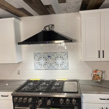 A stainless steel gas stove with a black hood vent centered below decorative floral backsplash tiles and white cabinetry.