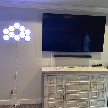 A wall-mounted TV with a soundbar above a light-colored wooden cabinet, with a hexagonal LED light display on the wall.