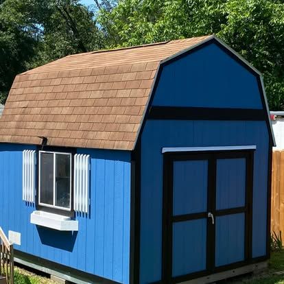 A blue shed with brown shingles and double doors, featuring a window with white shutters and a flower box, outdoors.