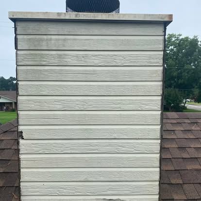 A chimney exterior covered in beige siding on a shingled roof, showing some staining and minor damage at the edges.
