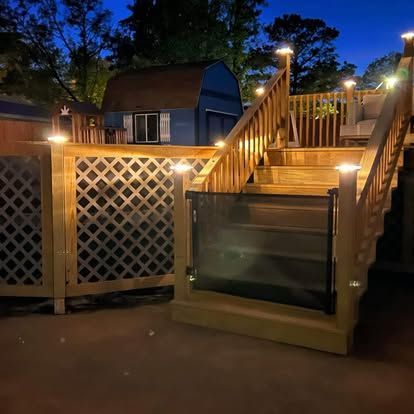 A wooden staircase at night, illuminated by small post cap lights, featuring a retractable black safety gate.