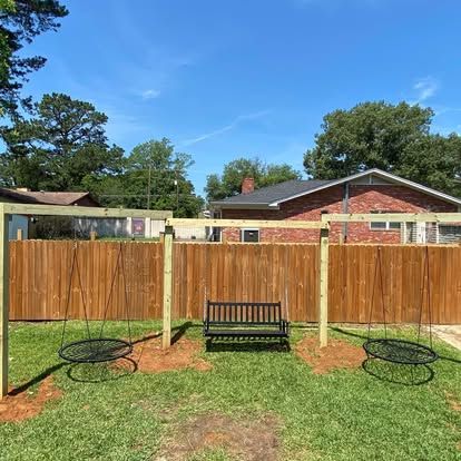 A wooden swing set frame in a grassy backyard, featuring two round platform swings and a central black bench seat.
