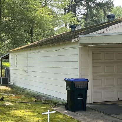 Side view of a white, horizontal-sided garage with two roof vents, a white door, and a black trash bin on a driveway.