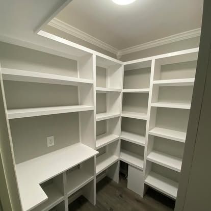 An L-shaped walk-in pantry with white built-in shelving against light grey walls and wood-look flooring.