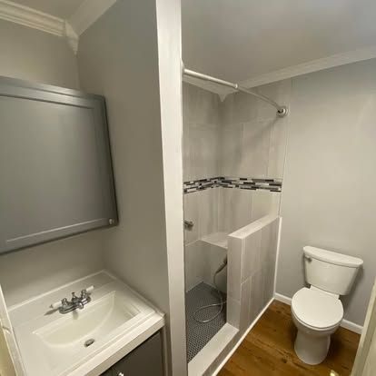 A bathroom with a gray vanity, white sink, a small walk-in shower with stone tile, and a white toilet on wood flooring.