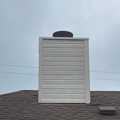 A white, horizontal-sided chimney chase topped with a dark metal chimney cap, positioned on a dark shingled roof.