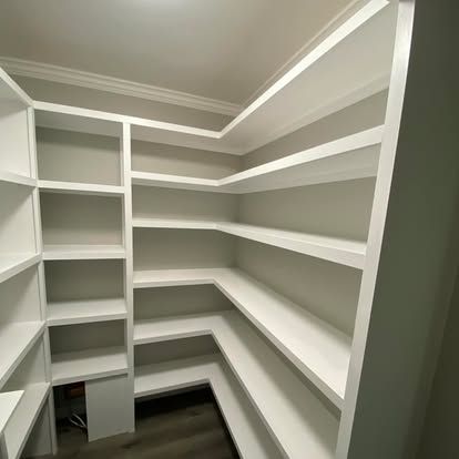 A corner pantry with white, custom-built shelving units against light grey walls and dark wood-look flooring.