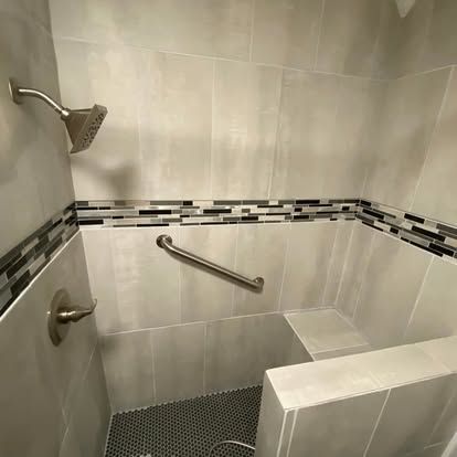 A walk-in shower with light gray tiled walls, a dark mosaic accent strip, a grab bar, a built-in seat, and dark floor tiles.