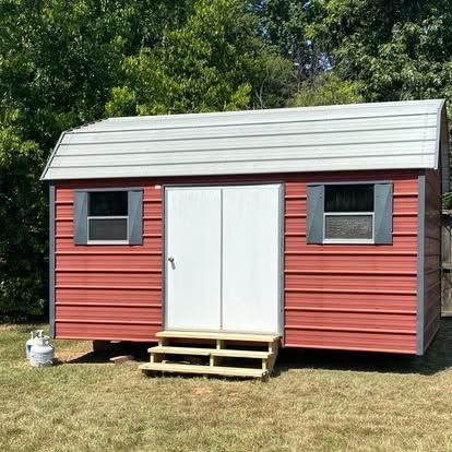 A red storage shed with a grey gambrel roof, white double doors, two windows, and wooden steps in a grassy yard.