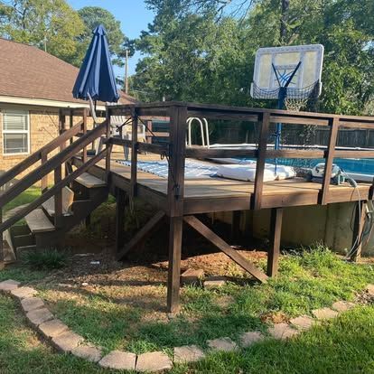A wooden deck with a staircase and a basketball hoop next to an above-ground swimming pool in a residential backyard.