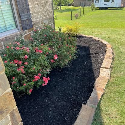 A stone-edged garden bed with pink rose bushes and dark mulch against a brick house wall.