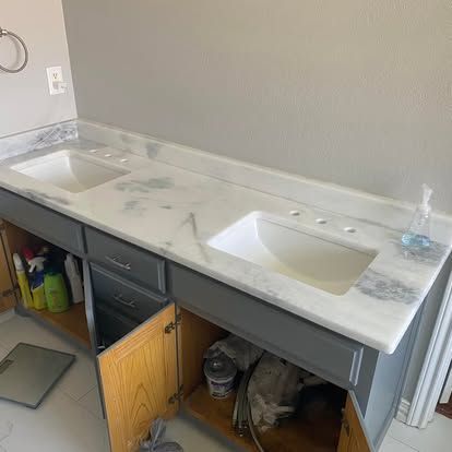 A double-sink bathroom vanity with a white marble countertop and gray cabinet doors, with one door left open.