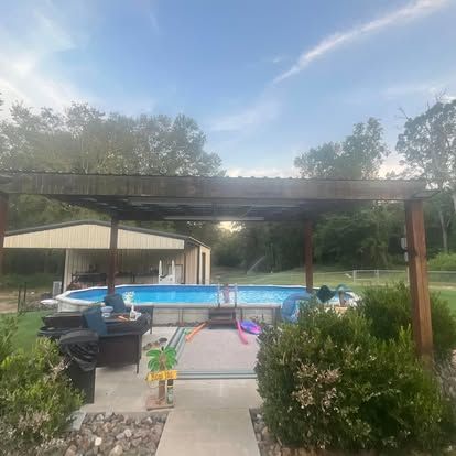 A backyard view featuring an above-ground pool under a wooden pergola, adjacent to a patio and an open-walled shed.