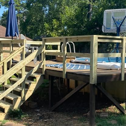 A wooden deck built around an above-ground pool, featuring stairs with railings and a nearby blue umbrella and basketball hoop.