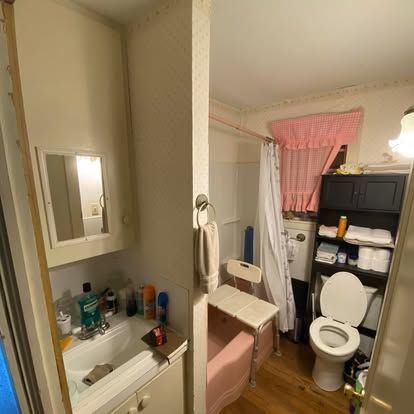 A bathroom with a white sink, a pink bathtub with a shower chair, and a toilet next to a black storage shelf.