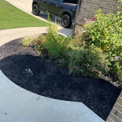 A curved landscaping bed with black mulch, green shrubs, and small plants next to a concrete path and a brick house wall.