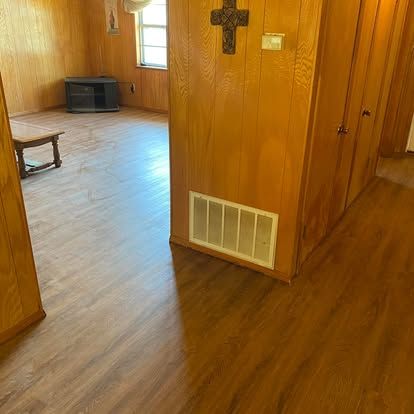 A wood-paneled room with light wood-look flooring, a wall-mounted cross, a floor vent, and a distant table and television.