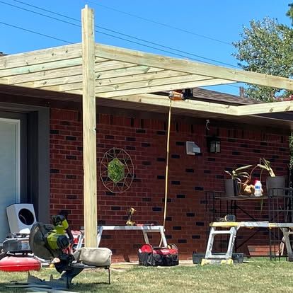 A wooden pergola structure under construction attached to a red brick house exterior with tools on the lawn.