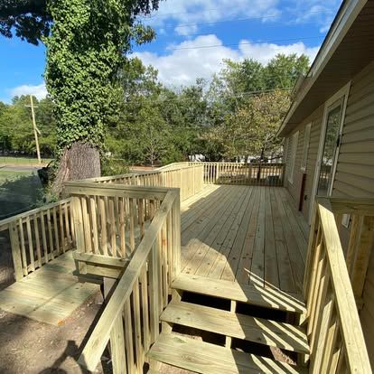 A newly constructed light wood deck with a ramp and stairs, extending from the side of a tan house near green trees.