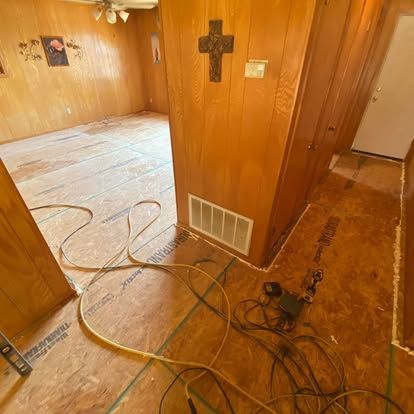 Interior room under renovation with bare plywood subfloors, wood-paneled walls, a wall-mounted cross, and scattered cables.