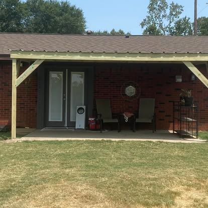 A covered concrete patio with two chairs and a small metal shelf attached to a red brick house under a brown roof.