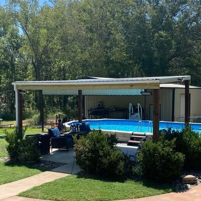 An above-ground swimming pool sits under a metal-roofed patio cover, surrounded by green trees and shrubs.