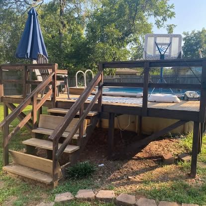 A raised wooden deck leading to an above-ground swimming pool with a basketball hoop, stairs, and a blue patio umbrella.
