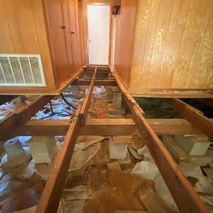 An under-floor crawl space showing exposed wooden joists, support blocks, and debris beneath a hallway.