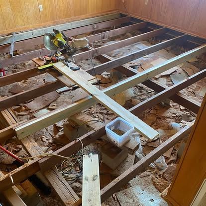 A room undergoing subfloor repairs with exposed floor joists, a miter saw, and new lumber beams resting on the framing.