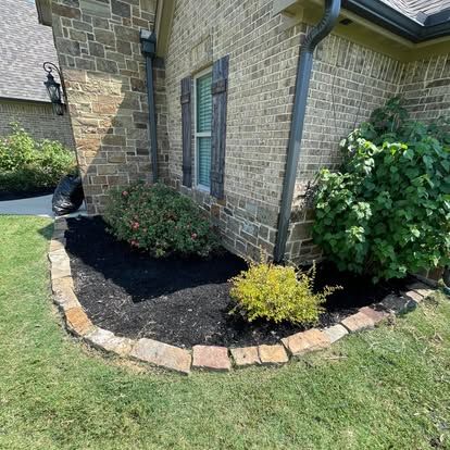 A brick house corner with a curved garden bed featuring dark mulch, small shrubs, and a stone border edge.