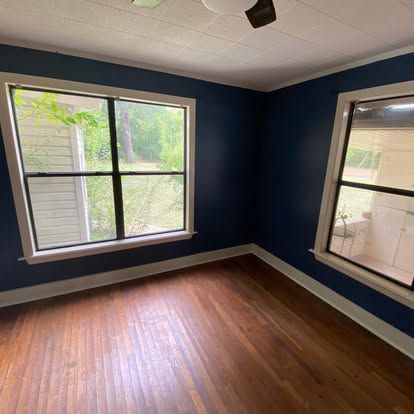 Empty bedroom with dark blue walls, hardwood floors, and two large windows looking outside.