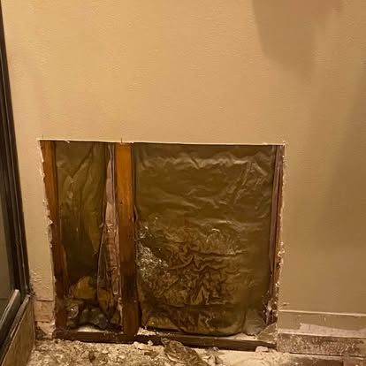 A rectangular hole in a beige wall reveals wooden studs and insulation behind the drywall.