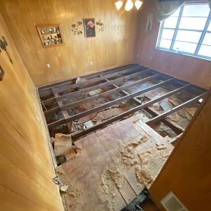A room with paneled walls under renovation, showing exposed floor joists and subflooring with debris.