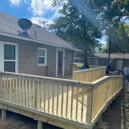 A newly constructed wooden deck with railings attached to the side of a brown house on a sunny day.