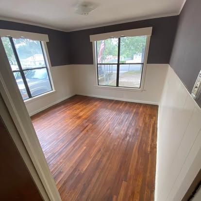 A vacant room with hardwood floors, white-painted lower walls, and dark-painted upper walls, featuring two windows.