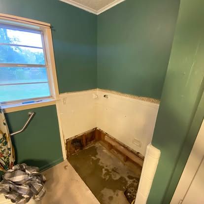 A small bathroom undergoing renovation with teal walls, exposed wall studs, and a bare concrete floor in the shower area.