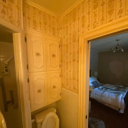 A bathroom with beige patterned wallpaper features a wall-mounted cabinet over a toilet, with a view into a bedroom.