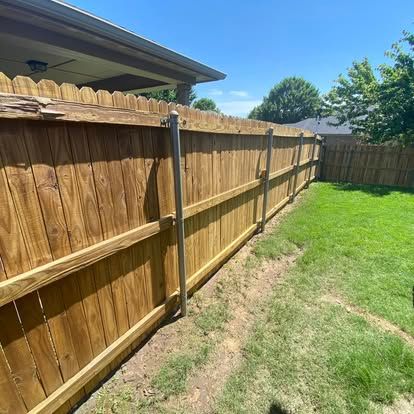 A wooden privacy fence with metal support posts, extending from a house along a grass lawn on a sunny day.