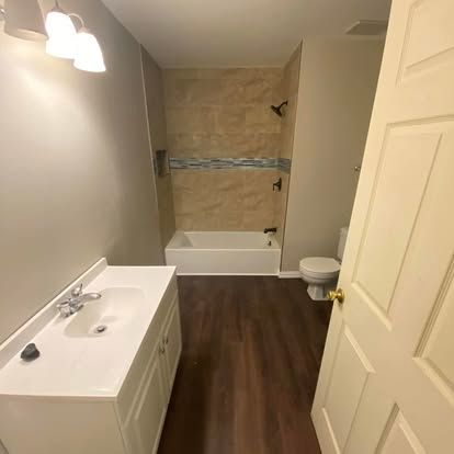 A bathroom with a white vanity, a bathtub with tan tile surround, and a toilet on a dark wood-look floor.