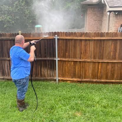 A person in a blue shirt uses a pressure washer to clean a dark wooden fence, revealing a lighter color underneath.