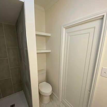 A bathroom showing a tiled shower, a toilet in a recessed alcove with open shelving, and a white closed door.