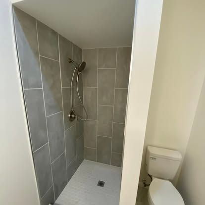 A small bathroom featuring a walk-in shower with grey rectangular tiles and a handheld sprayer next to a white toilet.