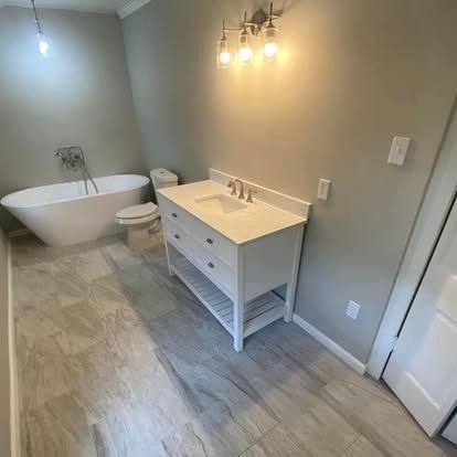 A modern bathroom featuring a white freestanding tub, a white vanity with a marble top, and gray wood-look tile flooring.