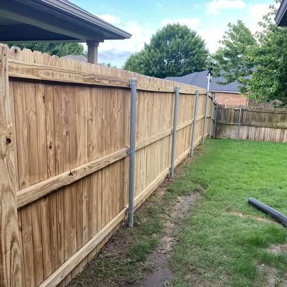 A wooden fence with metal support posts stands in a backyard with a green lawn and trees under a cloudy blue sky.