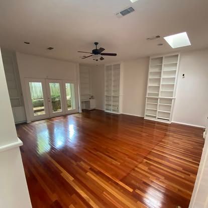 A spacious, empty room featuring rich, polished wood floors, white built-in bookshelves, and double glass patio doors.