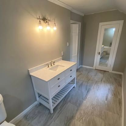 A modern bathroom featuring a white vanity with a basin sink, grey walls, wood-look tile flooring, and a glass-paneled door.
