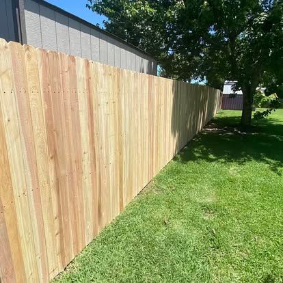 A tall, newly built wooden fence stretches across a green lawn with a tree and building in the background.